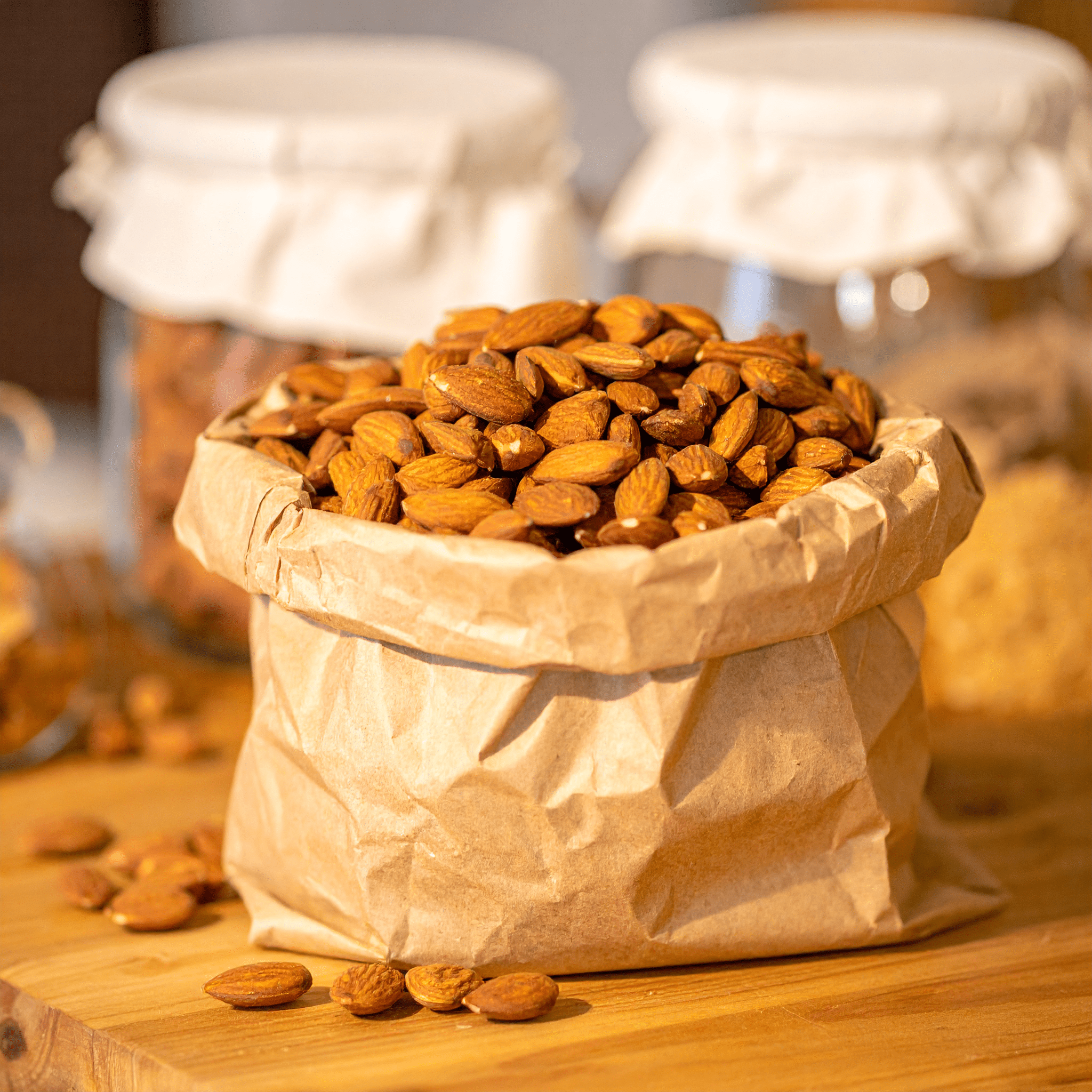Sac en papier rempli d’amandes posé sur une table en bois, avec des bocaux en verre floutés en arrière-plan.